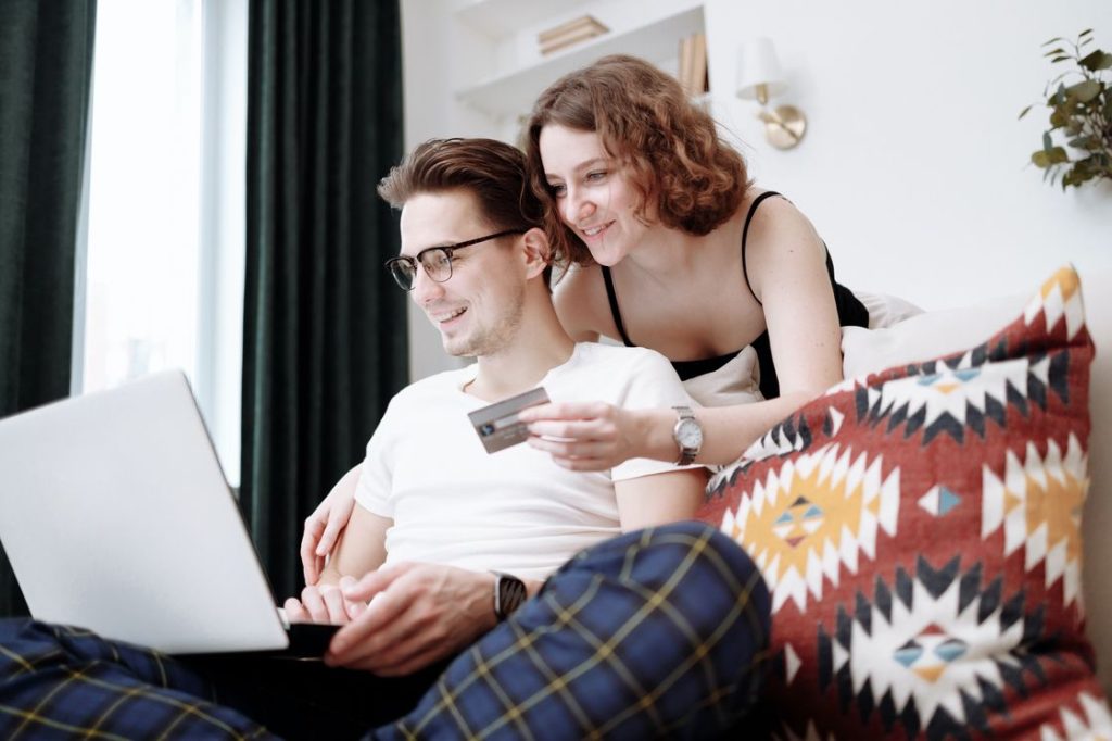 Couple looking at a laptop while the woman is holding a credit card and standing behind her husband