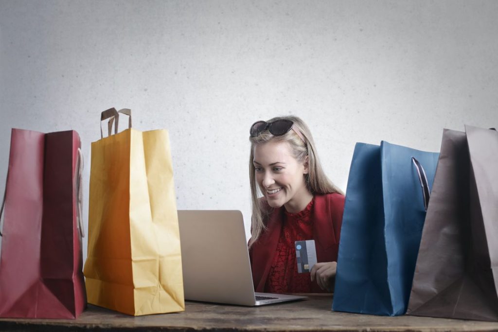 Woman holding her credit card while browsing through her laptop with multiple shopping bags placed around her