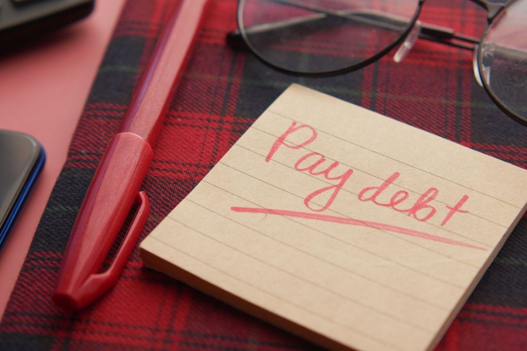 Notepad with a note written with "pay debt" beside a red pen and reading glasses