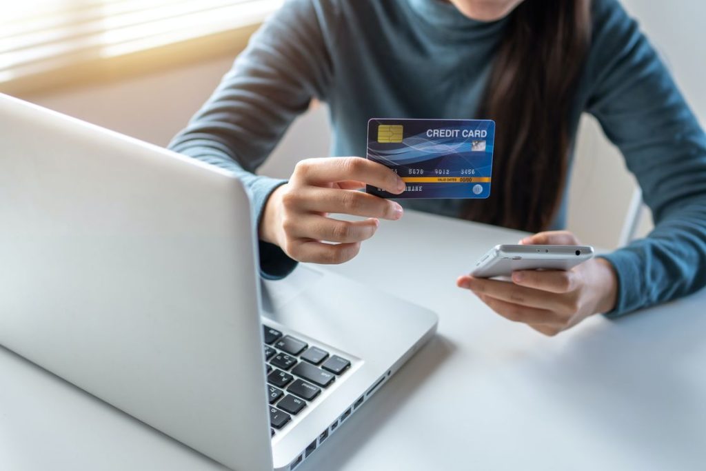 Woman holding out a credit card and a phone while using her laptop