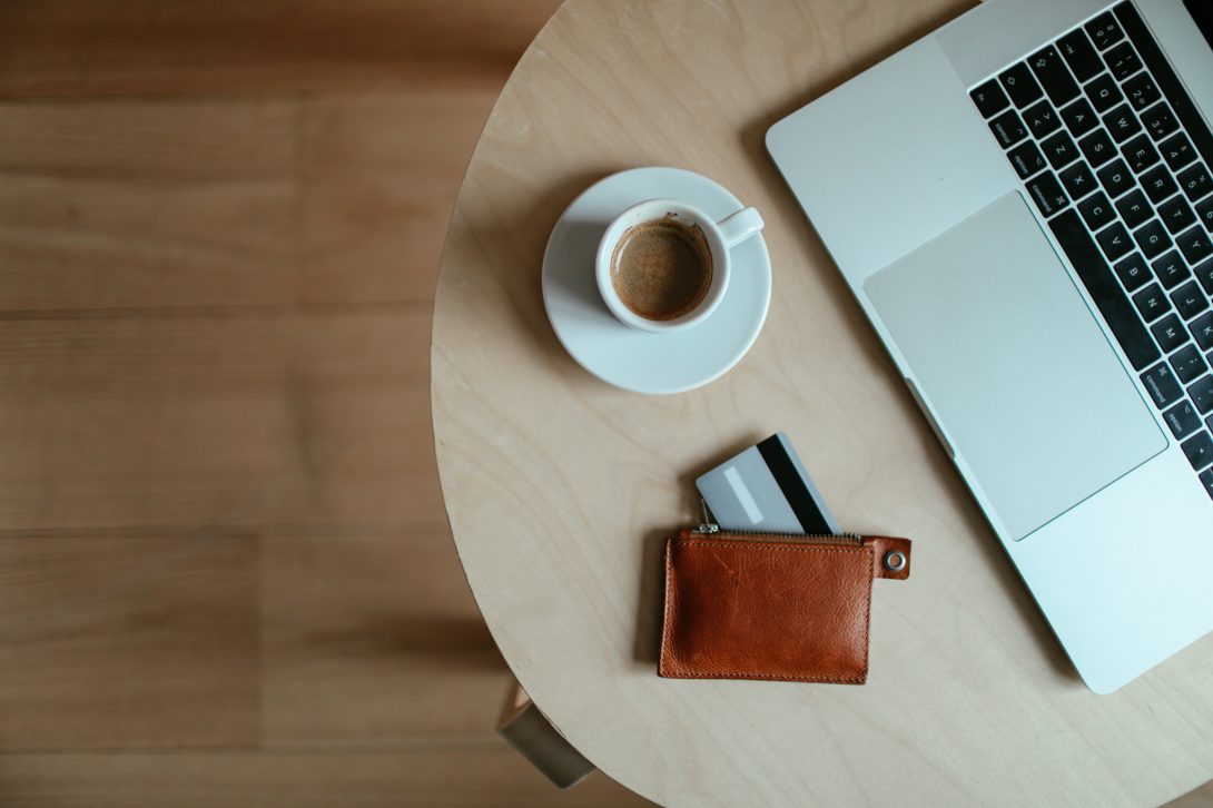 A cup of coffee on a saucer, laptop, and wallet with credit card placed on a table