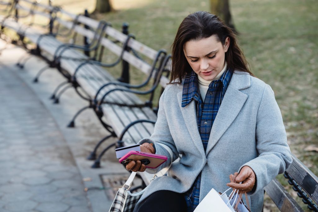 A woman sitting on a bench outside while holding her phone, credit card, and some shopping bags