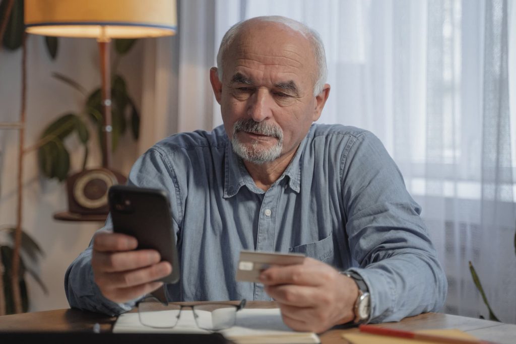 Elderly man holding both a smartphone and credit card while sitting at his table