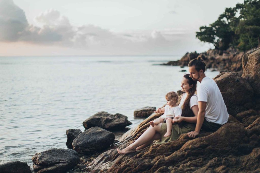 Couple with their baby daughter sitting on the rocky hill, enjoying the beach views