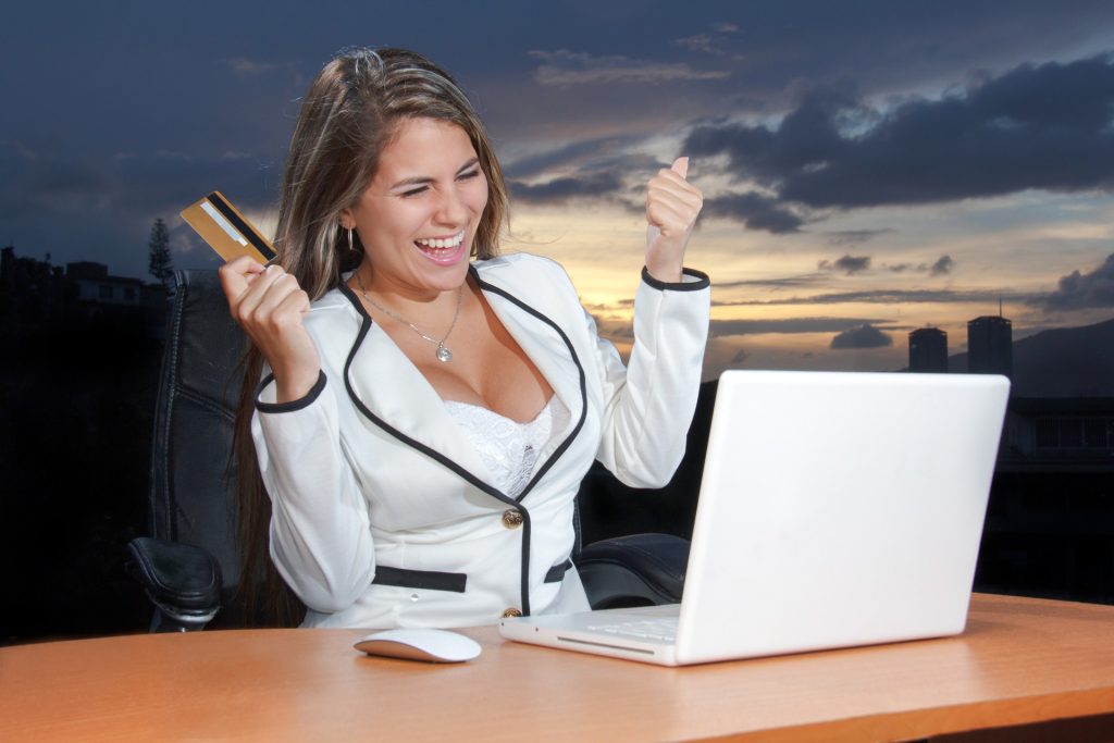 A woman sitting and smiling while holding a credit card in front of a white laptop