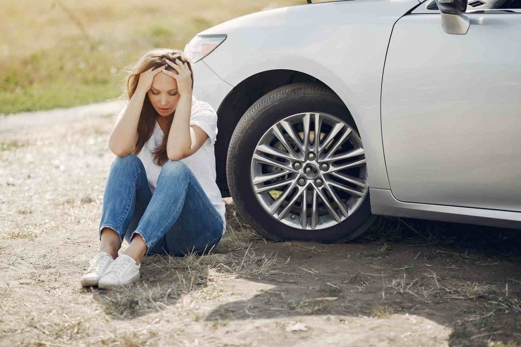 A woman sitting on the ground behind her car looking frustrated