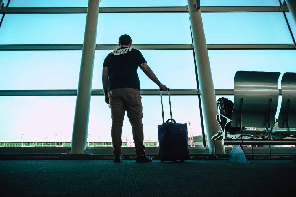 A man looking at glass window in the airport holding a rolling suitcase