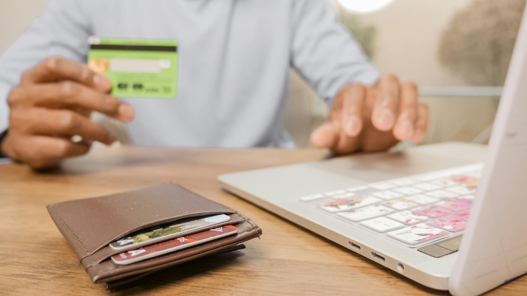 A man holding a credit card and working on a laptop, with a wallet on the table