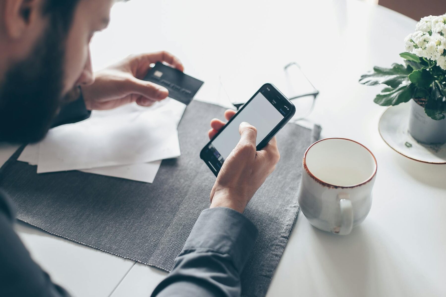 A man holding a credit card and using a smartphone with paperwork on the table