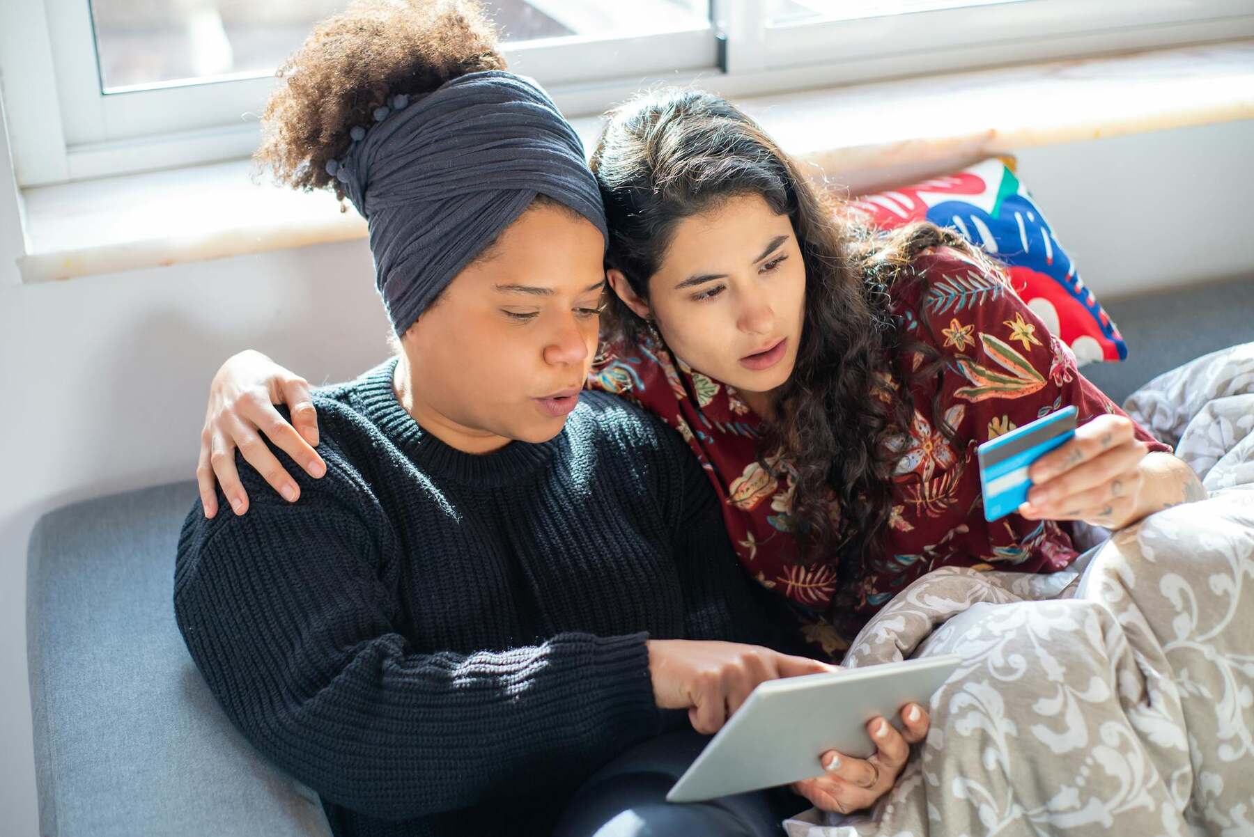 Two women on a couch, one showing a credit card to the other, while the other is pointing at her tablet