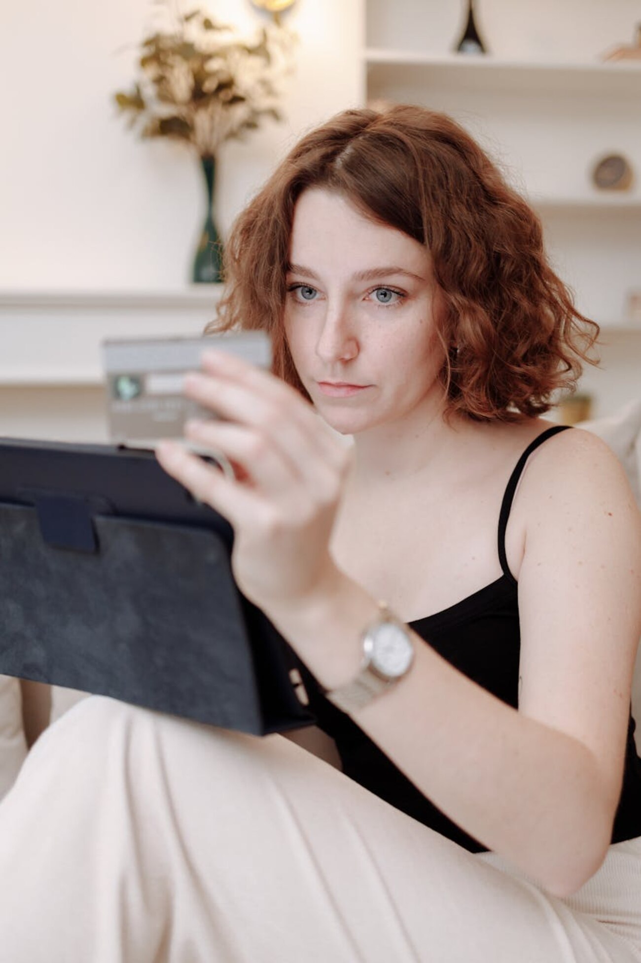 A woman sitting on a couch, holding a credit card while using a tablet