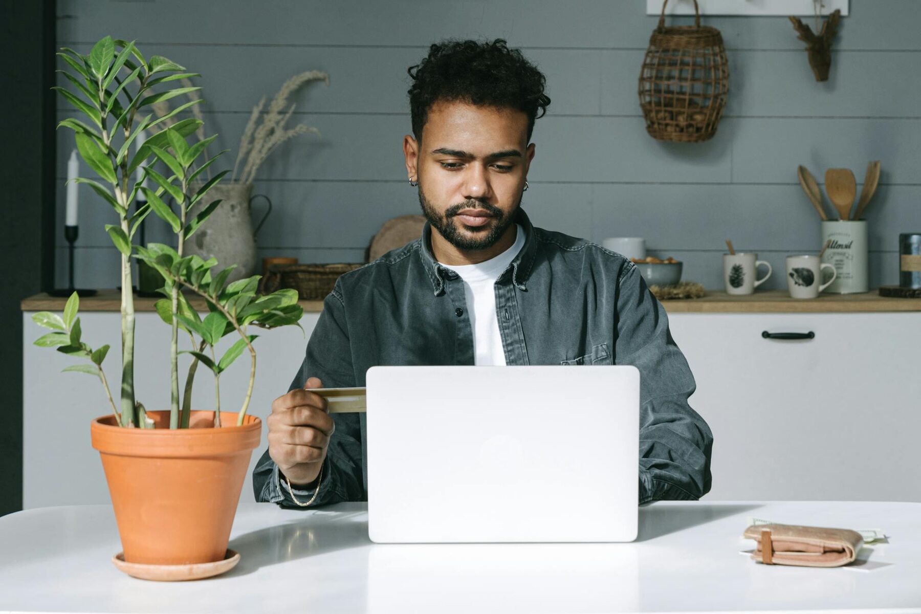 A man working on a laptop while holding a credit card at a table with a plant nearby