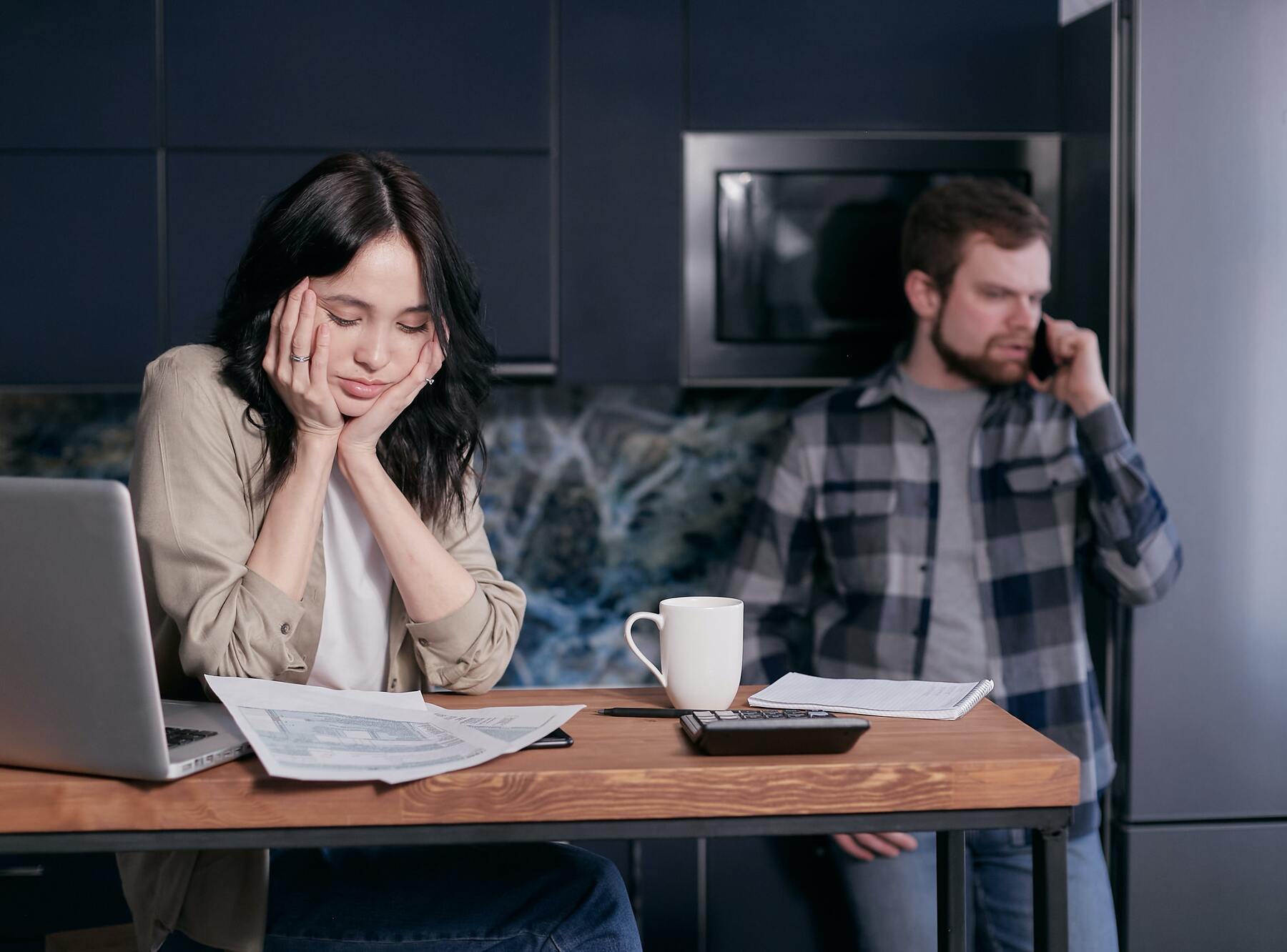 Woman looking distressed with the bills laid out on her table while her husband is on the phone