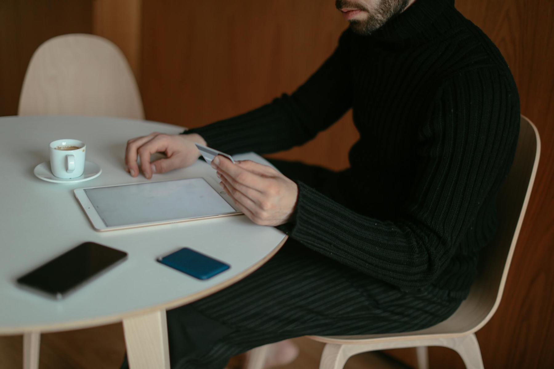 A man in a sweater sitting at a table, using a tablet and enjoying a cup of coffee