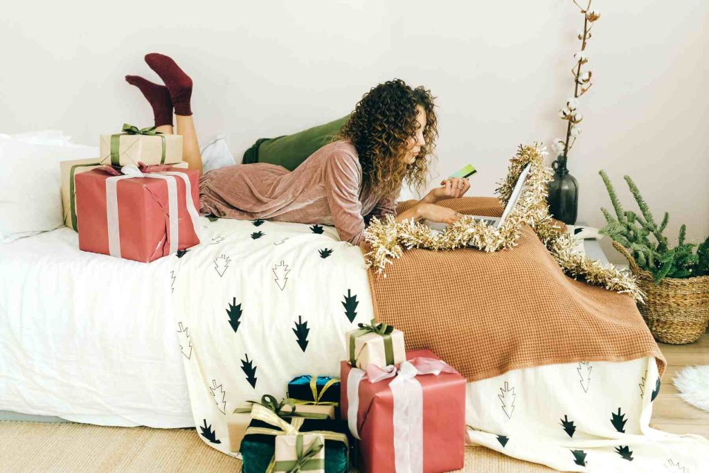 A woman on her bed using her laptop and holding her credit card with some shopping boxes on the ground