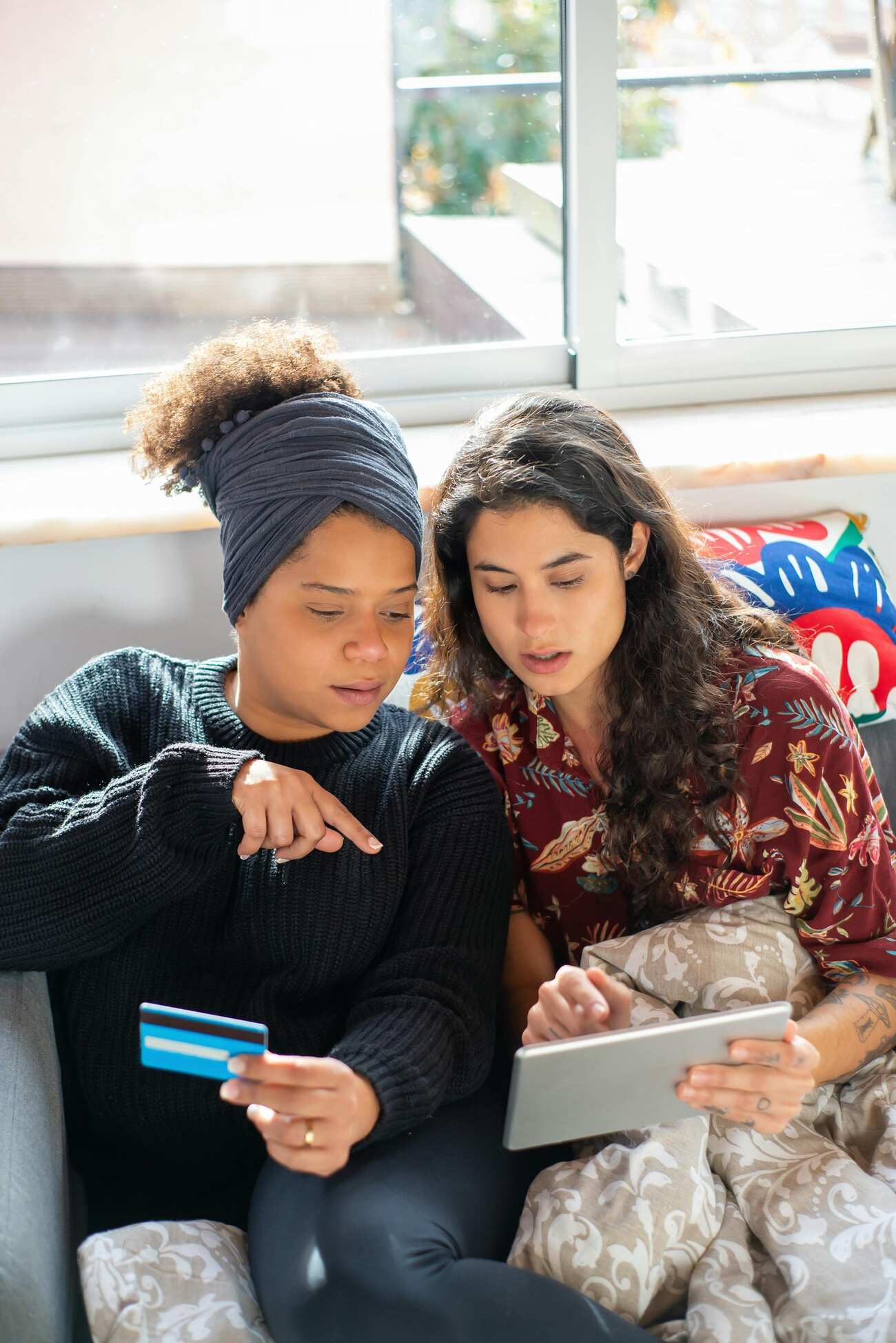 Two women sitting on a couch, one holding a credit card while the other holding a tablet