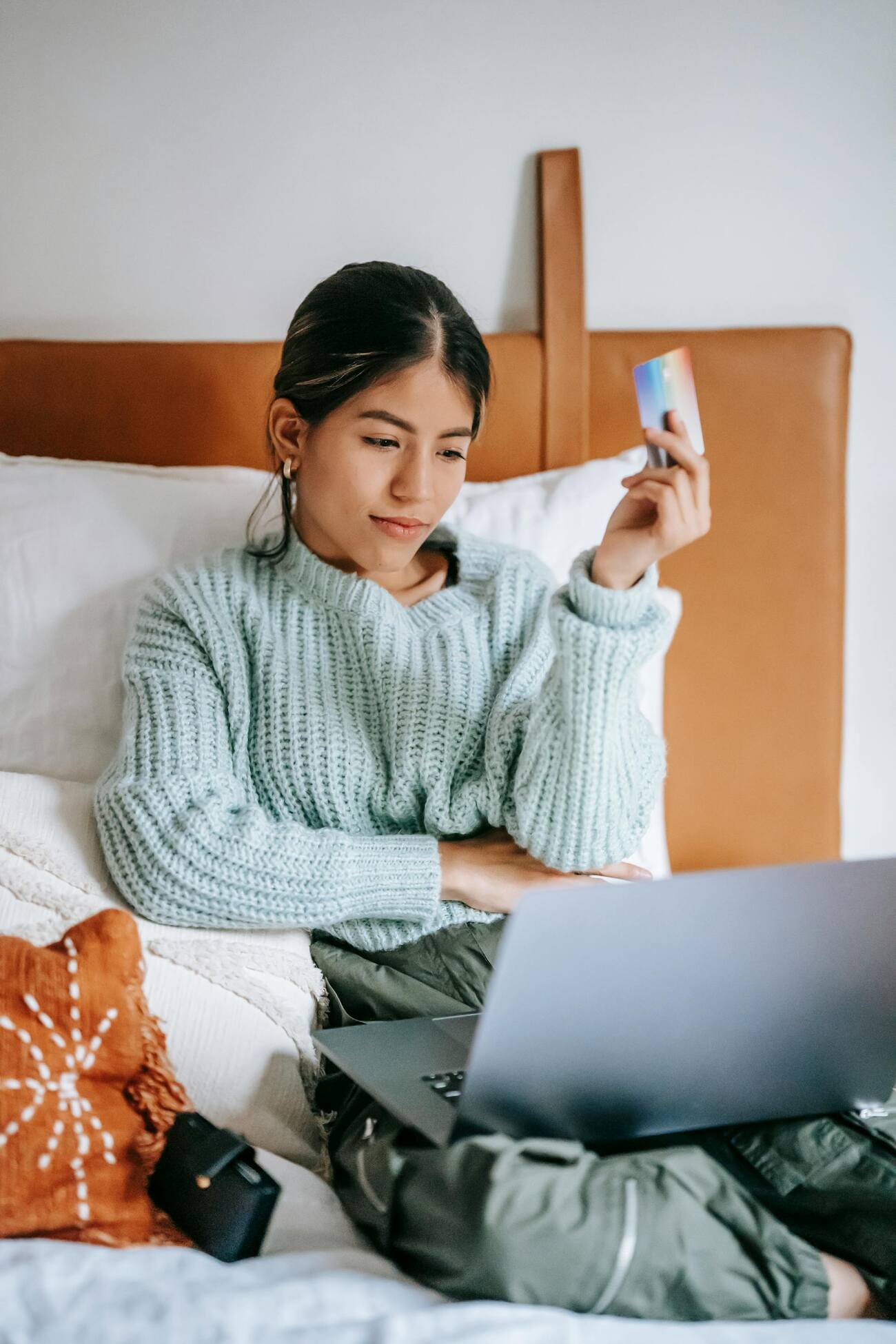 A woman sitting on a bed holding a credit card, engrossed in her laptop